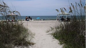 Un sentier qui longe la plage et mène à un belvédère surmonté d’un parasol bleu, le tout entouré de hautes herbes.