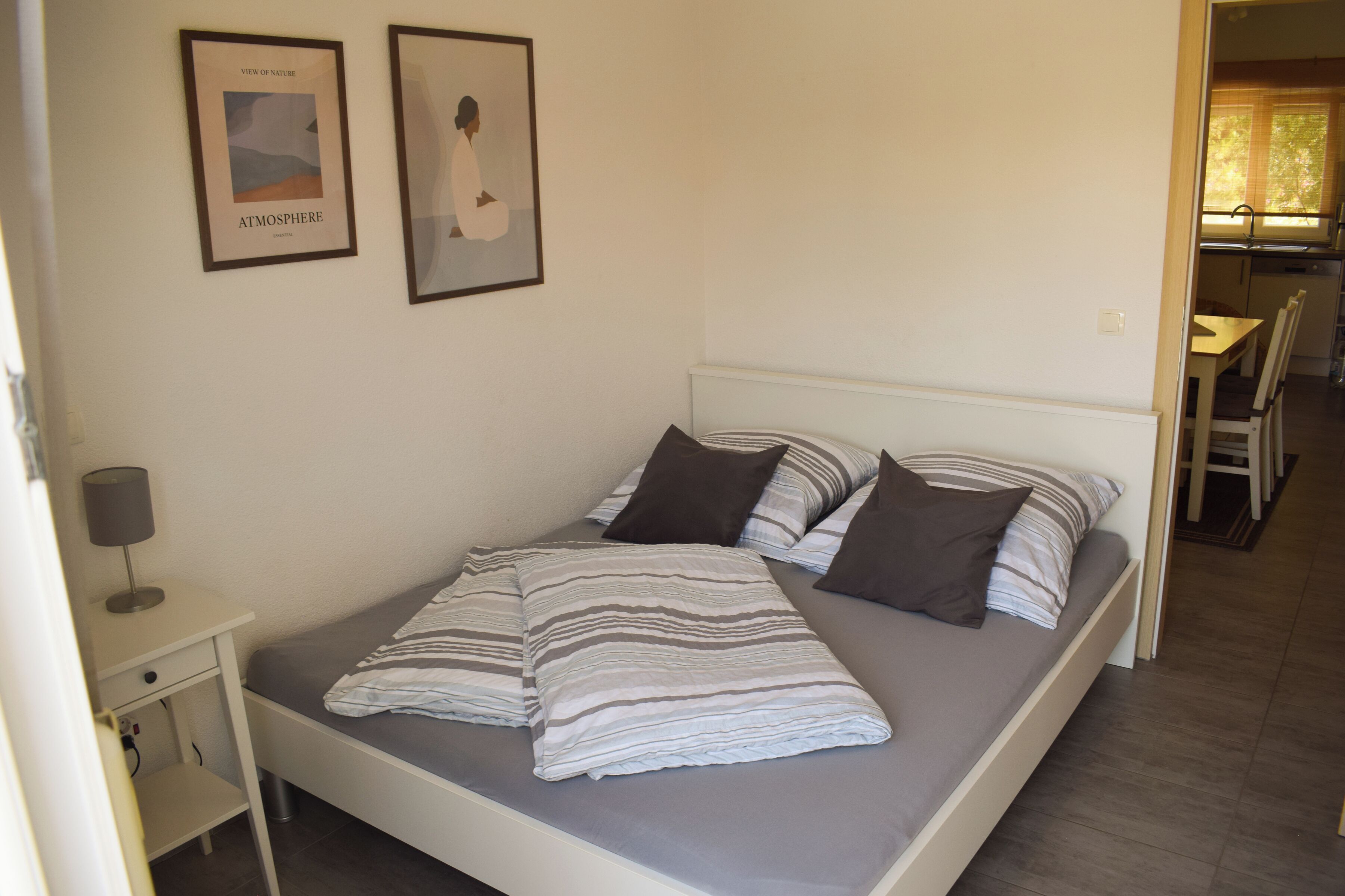A single bed with a gray mattress and striped bedding, a bedside table with a lamp, and two framed pictures on the wall.