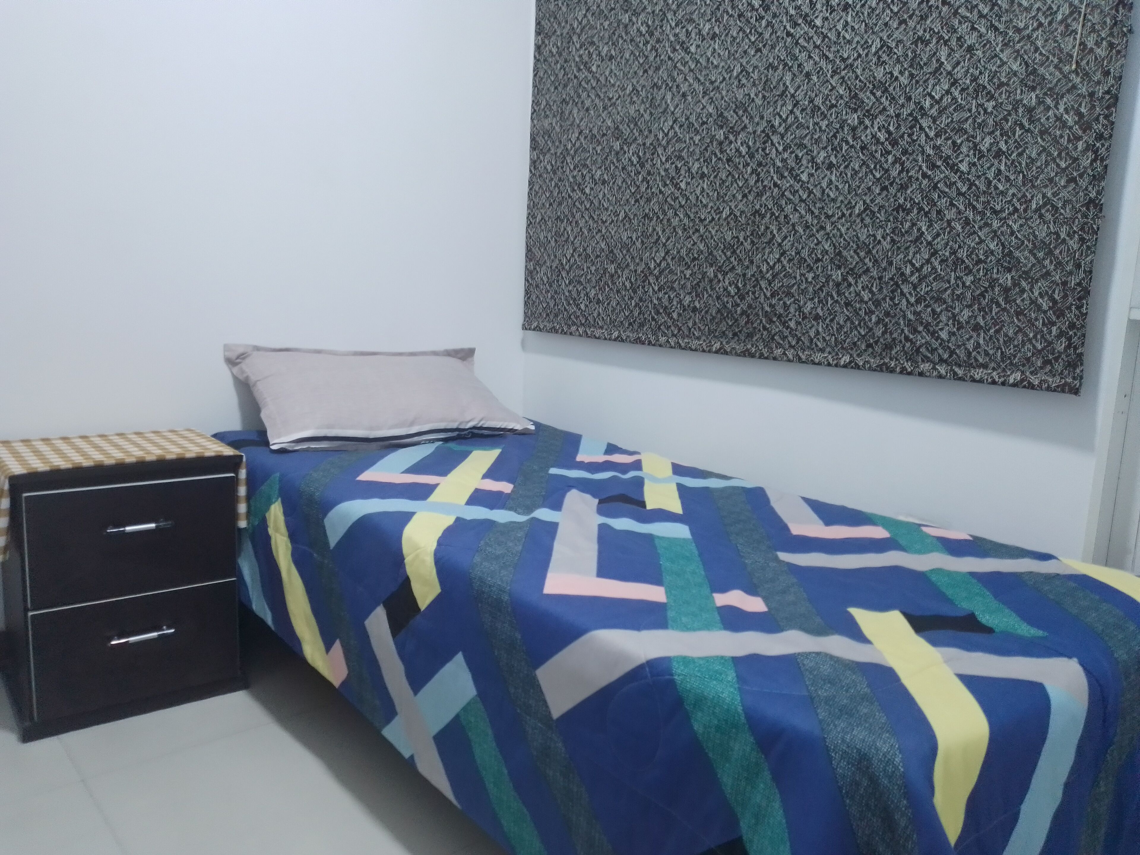 A small, neatly arranged bedroom with a bed, a bedside table with drawers, and a patterned wall hanging.