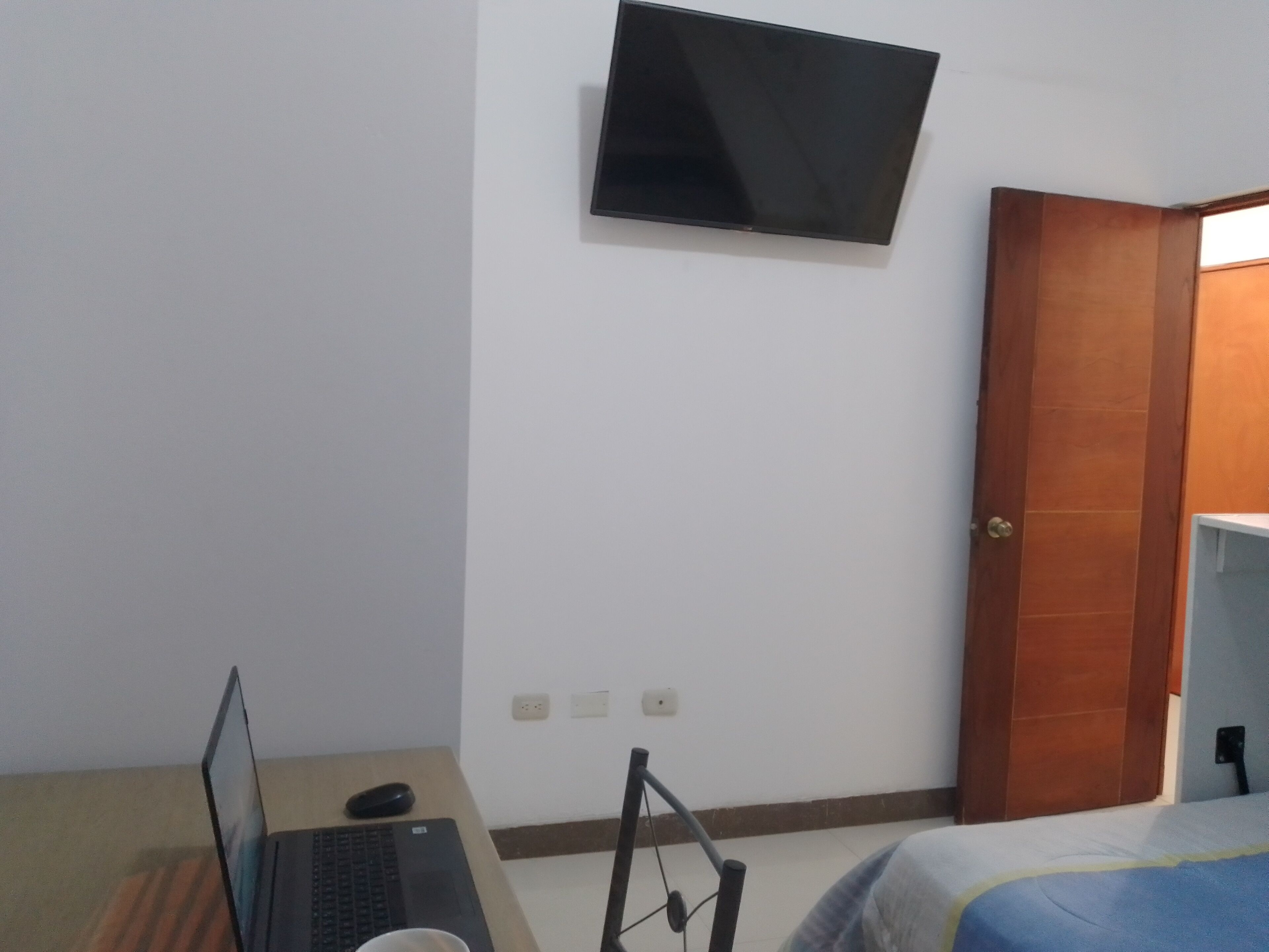 A hotel room with a bed, a desk with a laptop, a chair, a TV mounted on the wall, and a wooden door.