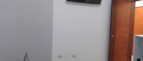A hotel room with a bed, a desk with a laptop, a chair, a TV mounted on the wall, and a wooden door.