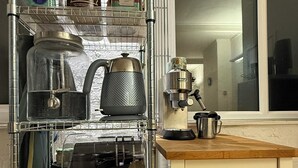 Una cocina con una estantería metálica que contiene diversos utensilios, una cafetera y una ventana con vistas.