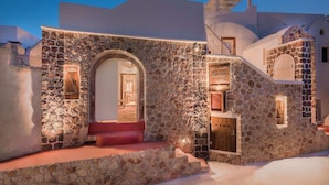 A stone building with a central arched doorway, a staircase leading to an upper level, and a small porch area. - Luxury Villas Hariton (Santorini)