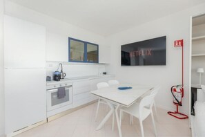 Eine moderne Küche mit weißen Schränken, einem eingebauten Ofen, einer Mikrowelle und einer Kaffeemaschine. Ein Esstisch mit weißen Stühlen und eine blaue Schüssel sind vorhanden. An der Wand ist ein Netflix-Schild zu sehen.