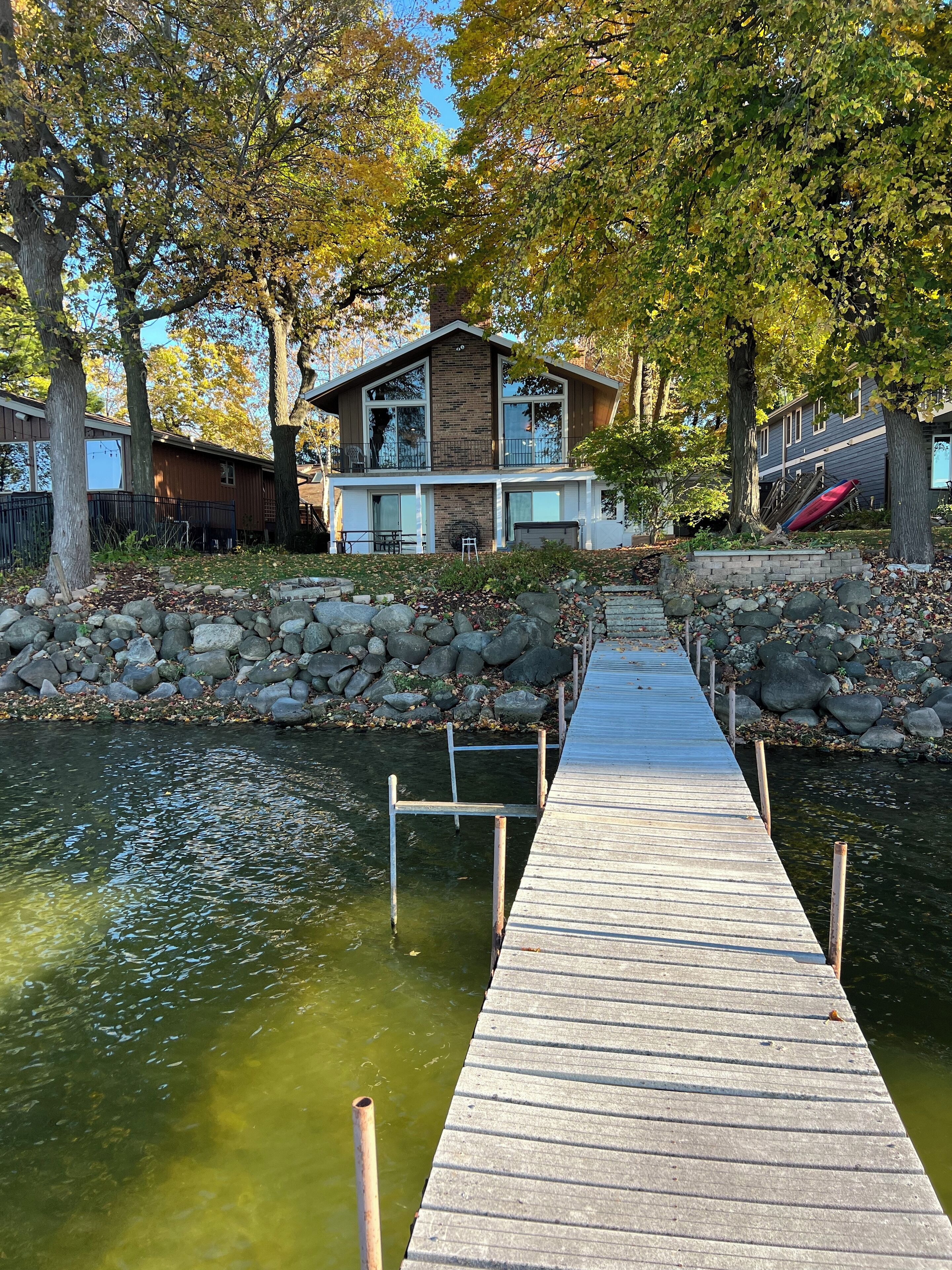 Une jetée en bois mène à une maison à la façade en briques, entourée d’arbres et bordée d’un rivage rocheux.