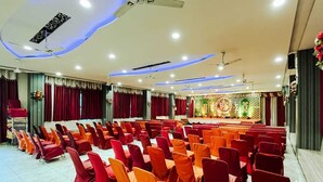 Une salle de conférence avec des rangées de chaises, une scène dotée d’un décor de fond et des ventilateurs de plafond.