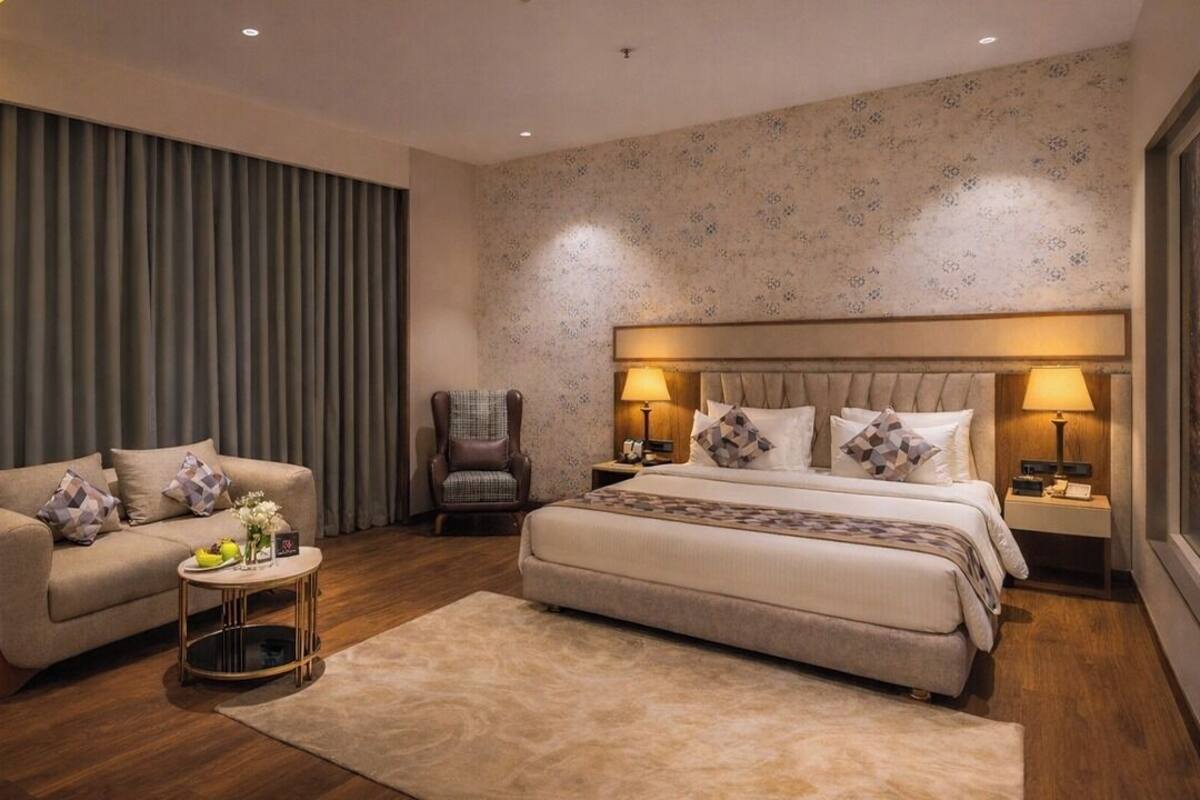 Una habitación de hotel con una cama grande, un sofá, una silla, una mesita redonda pequeña y una mesita de noche con lámparas.
