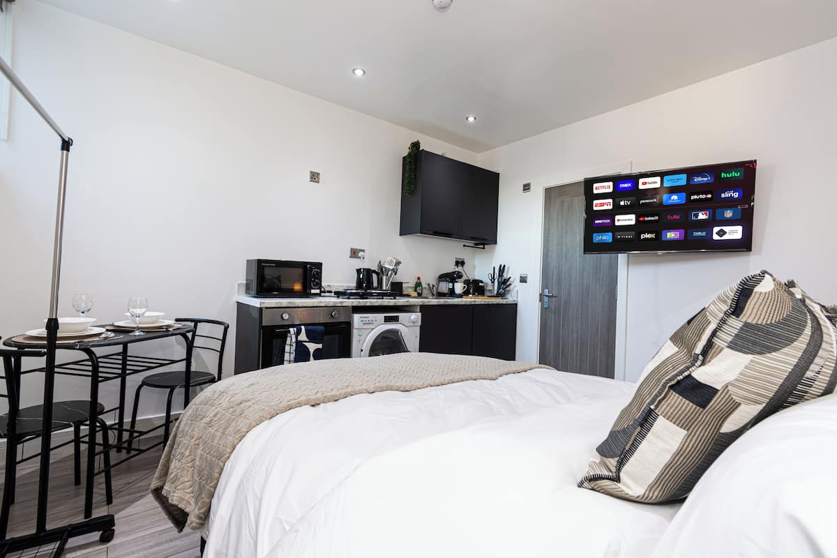 A compact hotel room with a bed, a small kitchenette, a flat-screen TV displaying streaming services, and a dining area with a table and chairs.