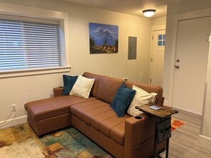 A brown leather sectional sofa with a matching ottoman, a small wooden side table, a colorful rug, a painting on the wall, and a window with blinds.