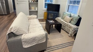 Ein modernes Wohnzimmer mit einem grauen Sofa, einem kleinen Couchtisch, einem Fernseher und einem Fenster mit Jalousien.