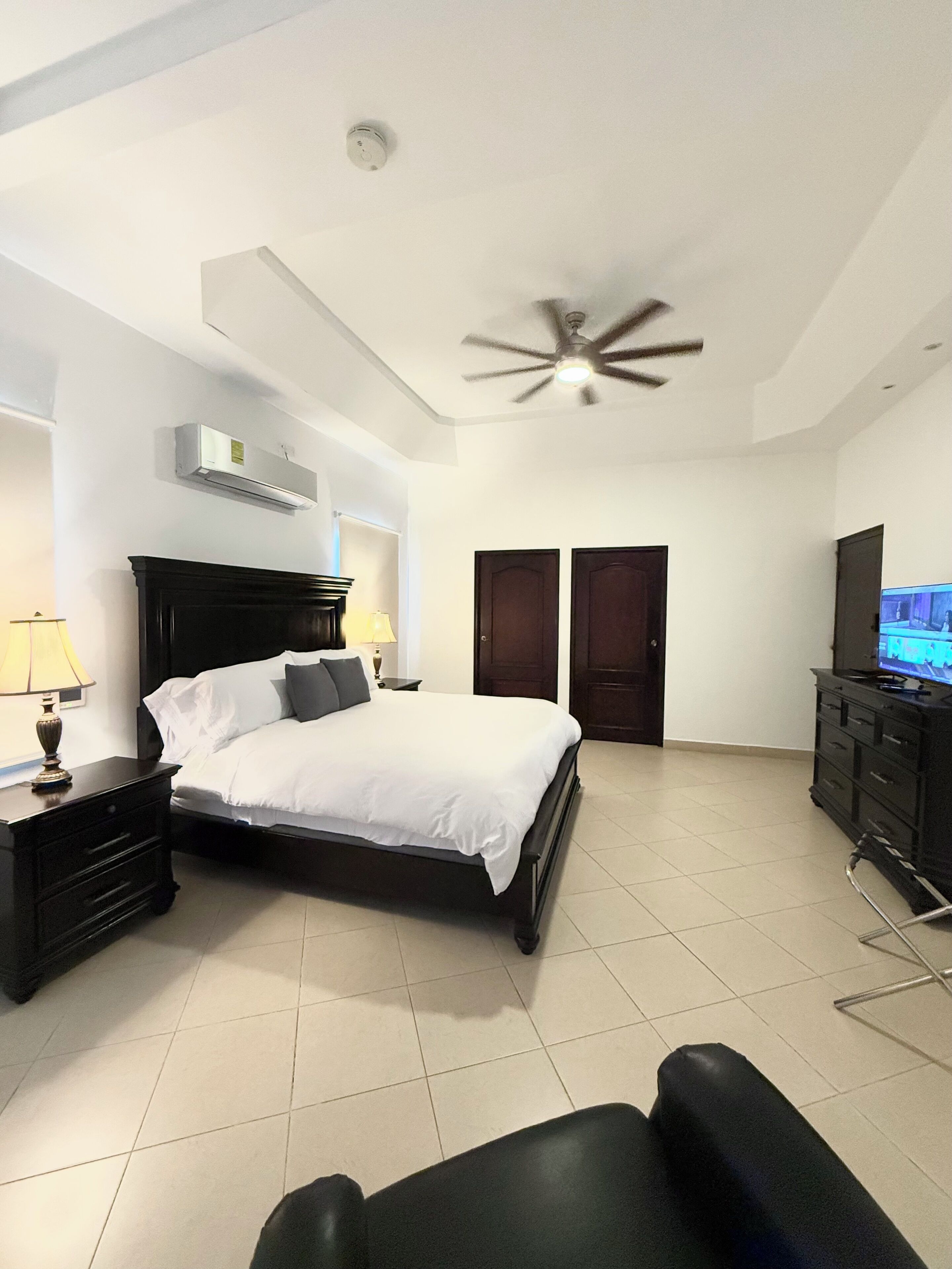 A bedroom with a bed, a ceiling fan, an air conditioning unit, a TV, and a dresser.