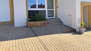 Een gebouw met een witte gevel, een trap die naar een deur leidt en een kleine plantenbak met groen.