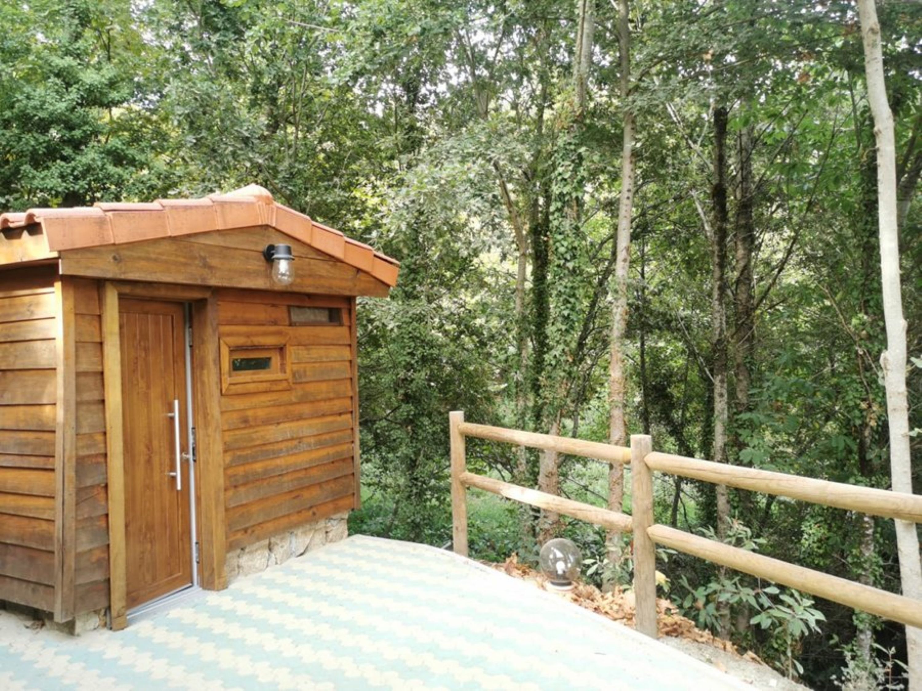 A wooden cabin with a door and a small window, surrounded by a wooden fence and dense forest.