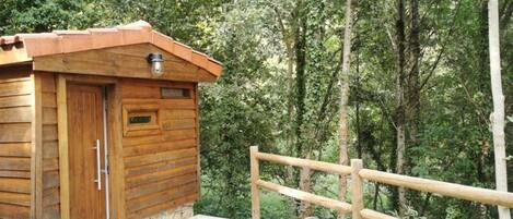 A wooden cabin with a door and a small window, surrounded by a wooden fence and dense forest.