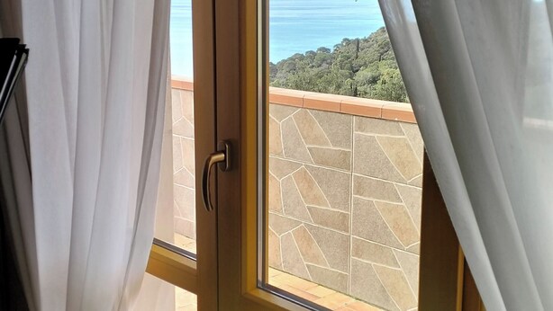A window with white curtains, a view of the sea, and a tiled patio area.