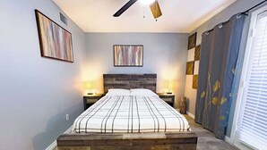 Une chambre à coucher avec une tête de lit en bois, deux tables de chevet avec des lampes, un ventilateur de plafond et une grande fenêtre avec des rideaux.