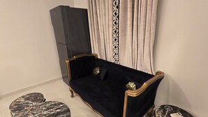 A dark blue sofa with gold trim, two marble-patterned side tables, and a black cabinet in a room with light-colored walls and curtains. - Karachi Beach Residency (Karachi)