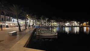 A nighttime waterfront scene with a docked boat, palm trees, and illuminated buildings along the promenade. - Island Paradise, peace and quiet with stunning views across to the lighthouse. (Platges de Fornells)
