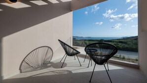 Un balcón con dos sillas de estructura metálica negra, vista a un paisaje con vegetación y una masa de agua, bajo un cielo despejado.