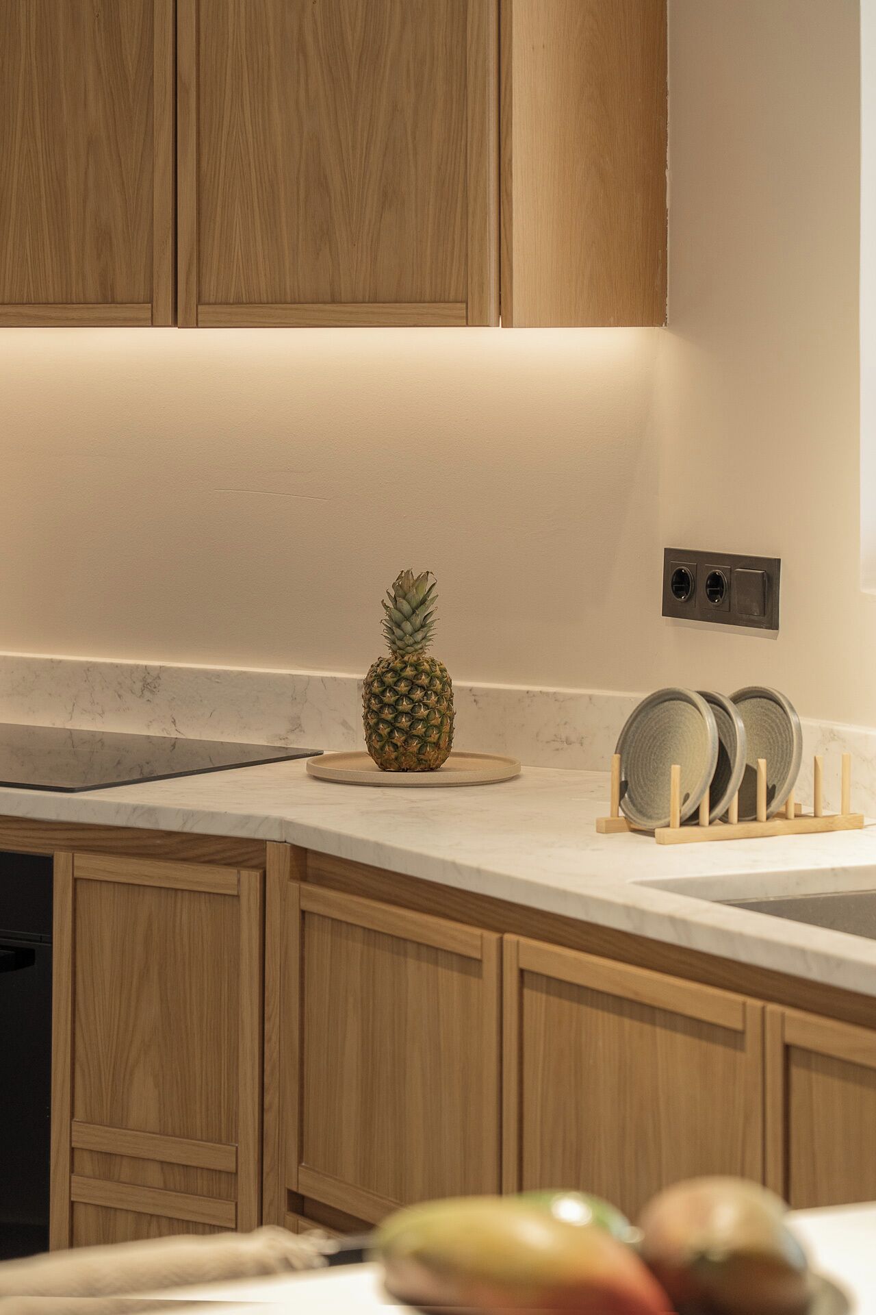 A kitchen with wooden cabinets, a marble countertop, a pineapple on a plate, and a dish rack with plates.