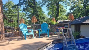 Un espace aménagé au bord de la piscine, avec une terrasse en bois, deux chaises bleues, un escalier pour accéder à la piscine et une zone délimitée par une clôture, où se trouvent des arbres.