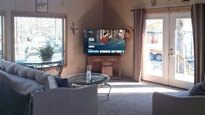 Un salon chaleureux avec une télévision, un plafond en bois et une vue sur l’extérieur par la porte.