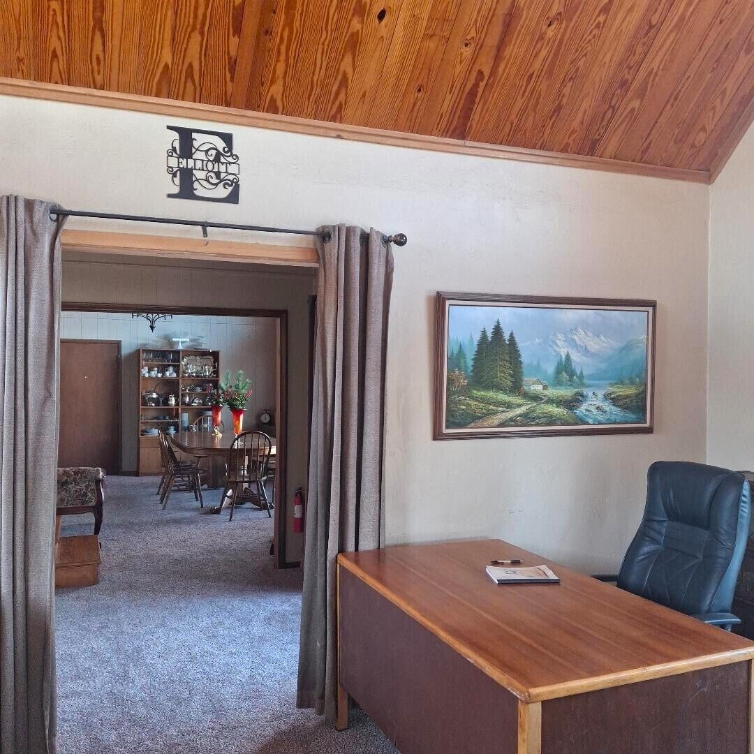 A room with a wooden ceiling, a desk, a chair, a bookshelf, and a painting of a landscape.