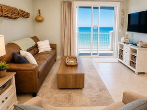 Ein Wohnzimmer mit braunem Ledersofa, einem Holztisch, einem weißen Schrank und Blick auf das Meer durch eine Glastür.