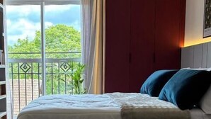 Une chambre à coucher avec un lit, un pouf vert, une chaise en bois et une vue sur la verdure par la fenêtre.