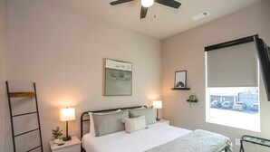Une chambre avec un lit, des tables de chevet, un ventilateur de plafond, une fenêtre donnant sur des voitures garées et un tableau accroché au mur.