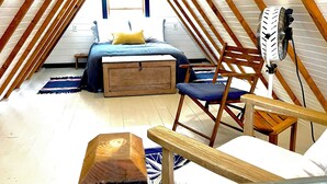 Slaapkamer in loftstijl met schuine wand, houten balken, een bed, een stoel, een ventilator en een klein tafeltje.