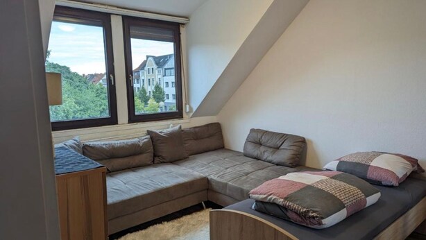A loft bedroom with a built-in sofa bed, a small wooden desk, and a large window offering a view of buildings and trees. - 2 room apartment (Bremen)