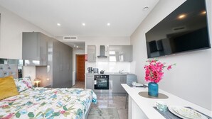 Une chambre moderne avec un lit, une table de chevet et une petite commode. On y trouve également une petite kitchenette équipée d’une cuisinière et d’un four. Un téléviseur est fixé au mur au-dessus de la kitchenette.