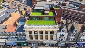 Un immeuble à plusieurs étages doté d’une terrasse sur le toit végétalisée, d’un style architectural classique avec de grandes fenêtres, et abritant divers commerces au rez-de-chaussée.