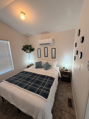 A bedroom with a large bed, an air conditioner, a nightstand with a lamp, and a wall-mounted clock.
