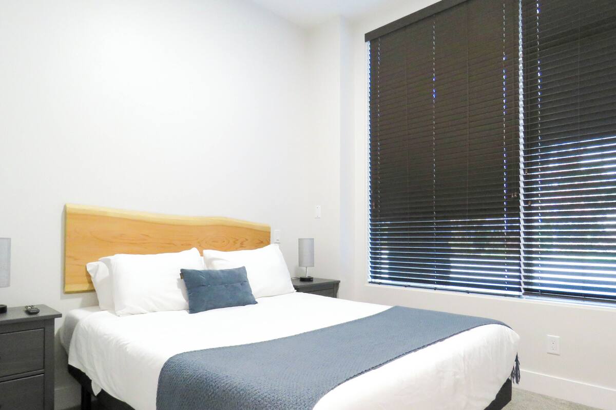 Cama bem arrumada com cabeceira de madeira, roupa de cama branca e manta azul. Tem uma mesa de cabeceira com uma luminária e uma tomada na parede.