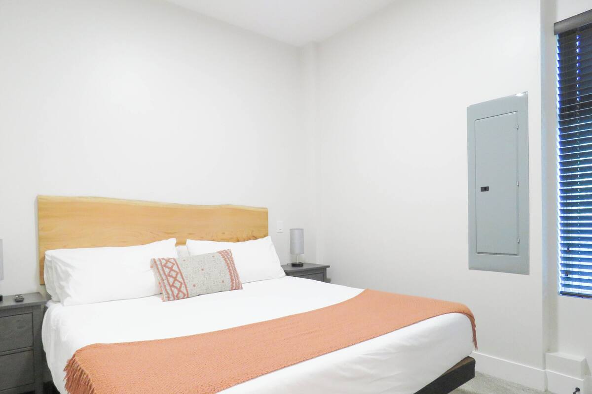 Quarto com cama, mesas de cabeceira, luminária na parede, porta e janela com persianas.