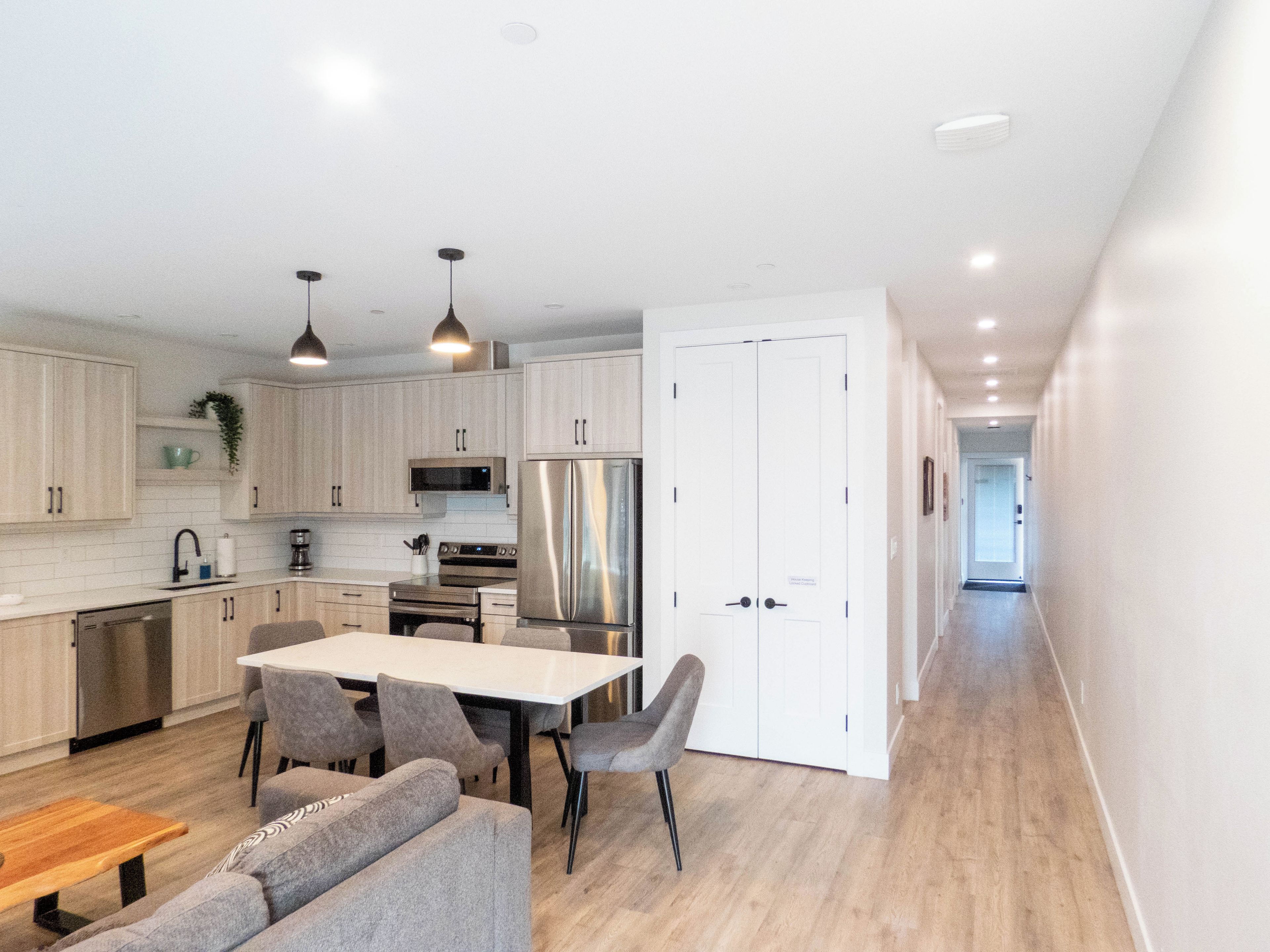 A modern kitchen with white cabinets, stainless steel appliances, a dining area with a table and chairs, and a hallway leading to another room.