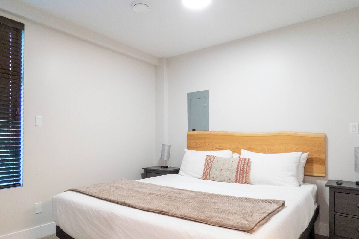 Quarto bem arrumado com uma cama de solteiro, mesas de cabeceira, um criado-mudo, uma luminária e uma janela com persianas.