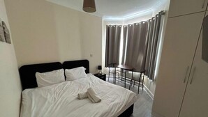 A hotel room with a bed, a nightstand, a table with chairs, a wardrobe, and a window with curtains. - Gassiot House (London)
