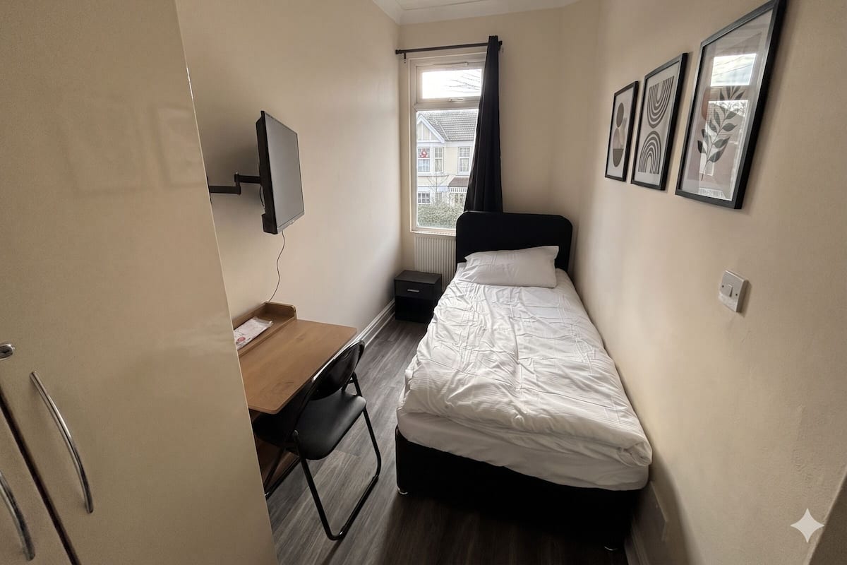A small, compact room with a bed, a desk, a chair, a TV mounted on the wall, and framed pictures on the wall.