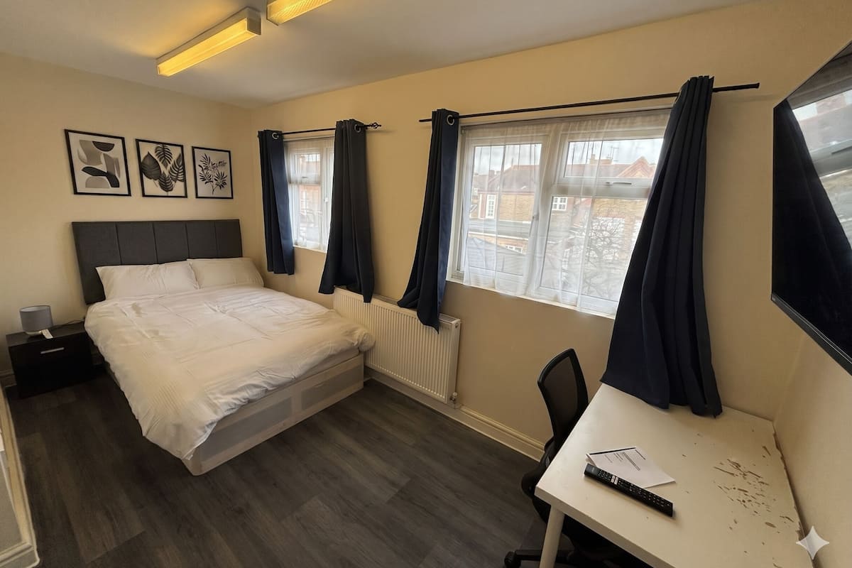 A hotel room with a bed, a desk, a chair, a radiator, and a window with curtains.