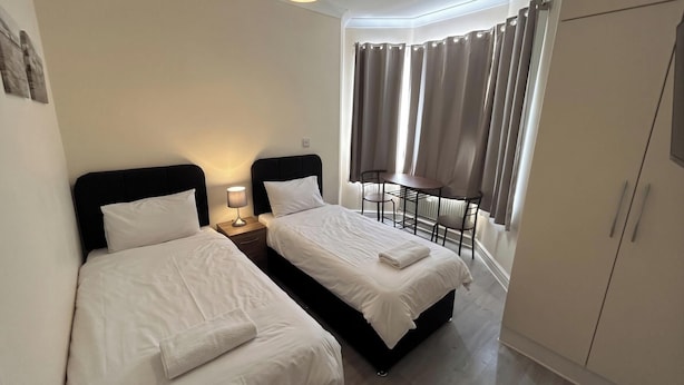 A hotel room with two beds, a desk, a chair, a wardrobe, and a window with curtains. - Gassiot House (London)