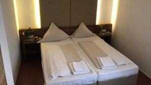 A double bed with two pillows and a folded towel, a nightstand with a vase and a fruit bowl, and a headboard with a light fixture. - Hotel Riede (Bremen)