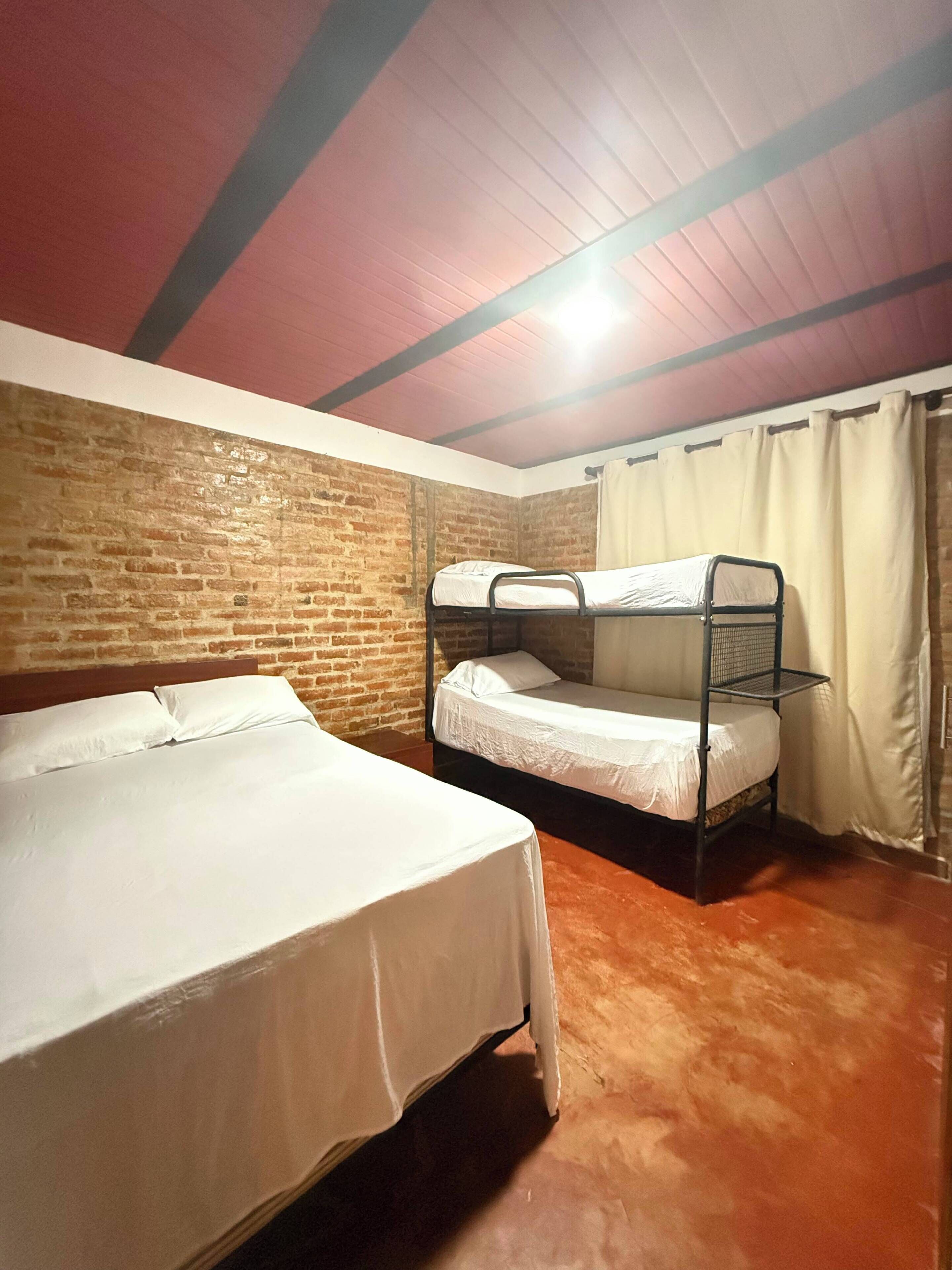 A room with a bunk bed and a single bed, both with white bedding. The room has a brick wall and a wooden ceiling with exposed beams.