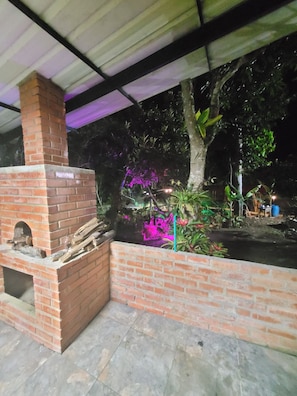 Un horno de ladrillo al aire libre bajo una estructura cubierta, con suelo de baldosas, rodeado de vegetación y una pequeña mesa al fondo.