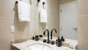 Un baño con encimera de granito, un lavabo blanco, grifos negros y un espejo. Hay dos toallas blancas colgadas en ganchos sobre el espejo.