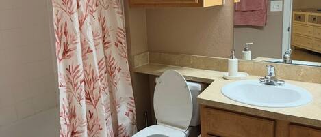 Et badeværelse med toilet, vask, træskabe, badeforhæng og et spejl.