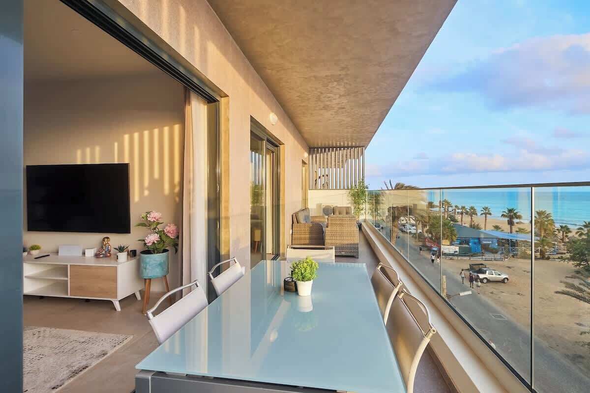 Una sala de estar moderna con una mesa de vidrio, un televisor de pantalla plana y un balcón con vista a la playa.