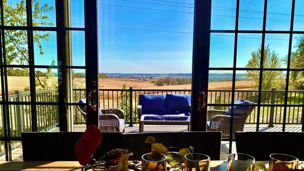 A room with large windows overlooking a countryside view, furnished with wicker chairs and a table set with candles and flowers.